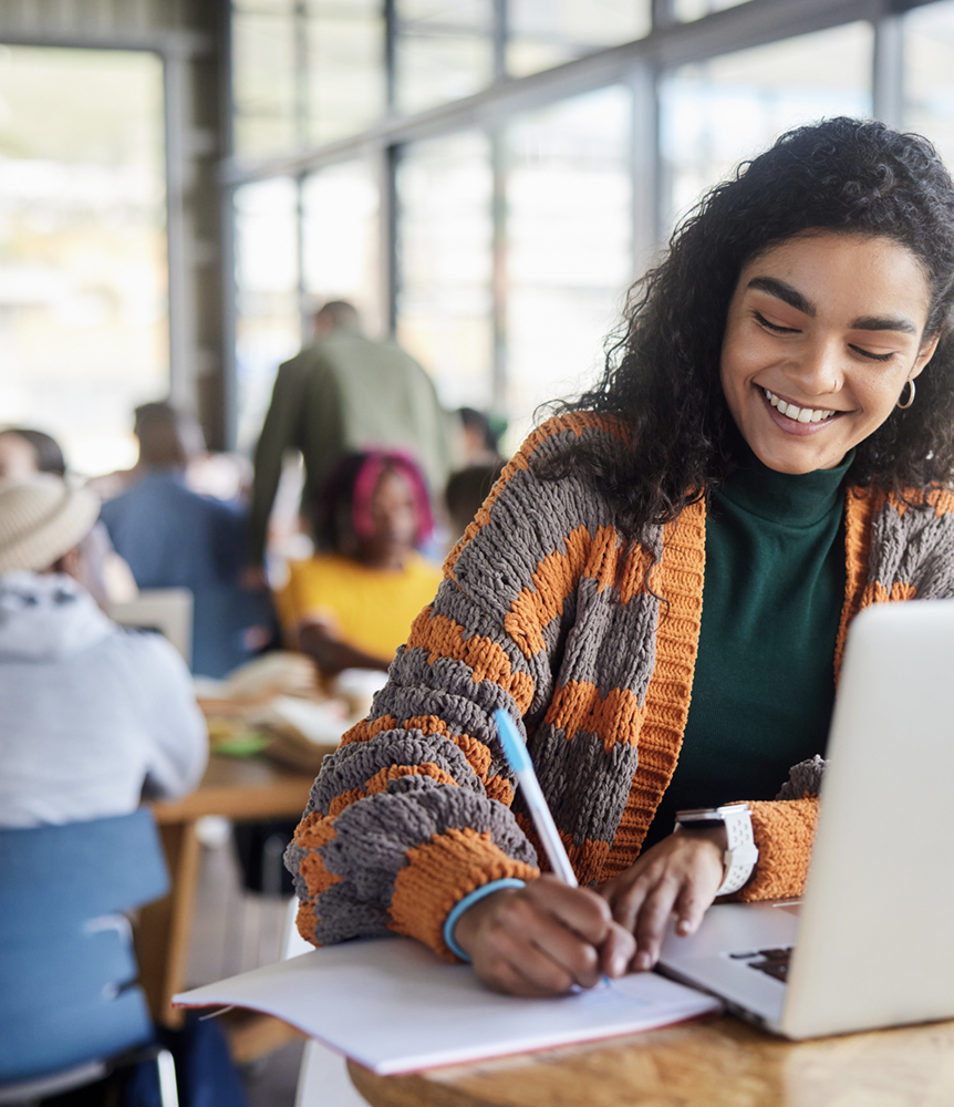Smiling community college student writing notes beside her laptop in a busy classroom